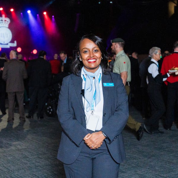 A woman in a professional suit smiles confidently, standing amidst a lively event with guests chatting and a stage lit with colorful lights. The Te Pae branding is visible on her name tag.